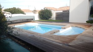 Une piscine dotée d’une terrasse en bois, entourée d’un mur blanc et d’un petit jardin.