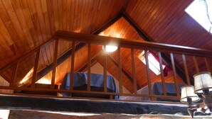 A loft bedroom with a wooden ceiling and a sloped roof. There is a bed with a blue cover and a lamp on a table.