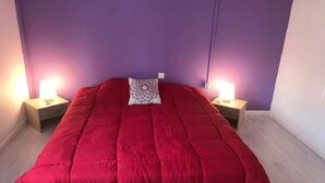 Cama com colcha vermelha, duas luminárias de cabeceira e parede roxa.