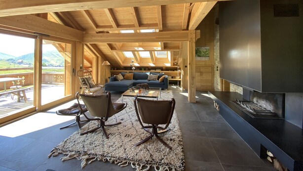 Ein großzügiger Wohnbereich mit Holzbalkendecke, einem blauen Sofa, zwei Sesseln, einem Kamin und Blick ins Freie.