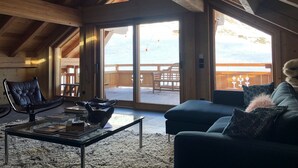 Ein gemütliches Wohnzimmer mit Holzbalkendecke, einer Glastür, die auf einen Balkon führt, einem Ledersessel, einer Couch mit Zierkissen und einem Couchtisch mit Büchern.