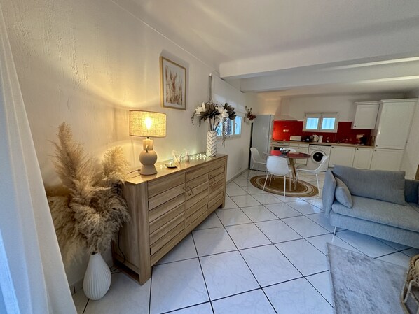 Een woonkamer met een bank, een houten kast en een eettafel. Op de achtergrond een keuken met rode kastjes en een koelkast.