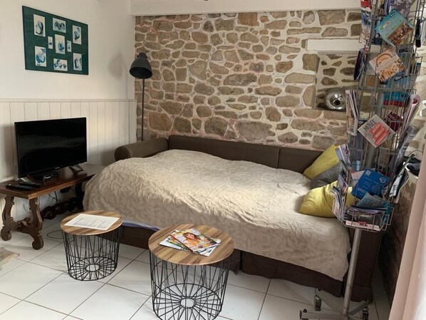 A cozy room with a stone wall, a bed with a beige blanket, a TV on a wooden table, and two small round tables with magazines.