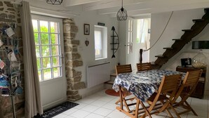 Un coin repas avec une table recouverte d’une nappe à motifs, des chaises en bois, un mur en pierre, une fenêtre avec des rideaux et un escalier.