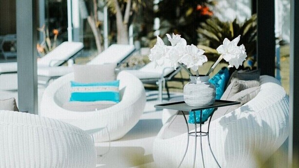 Un moderno espacio lounge al aire libre con asientos blancos acolchados, una mesita con un jarrón de flores blancas y vistas a una piscina y follaje.