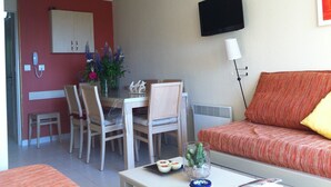 A room with a dining table, chairs, a sofa, a TV mounted on the wall, and a telephone on the wall. - Apartment, facing the sea, near the beach, 6 people, 2 bedrooms (Le Lavandou)