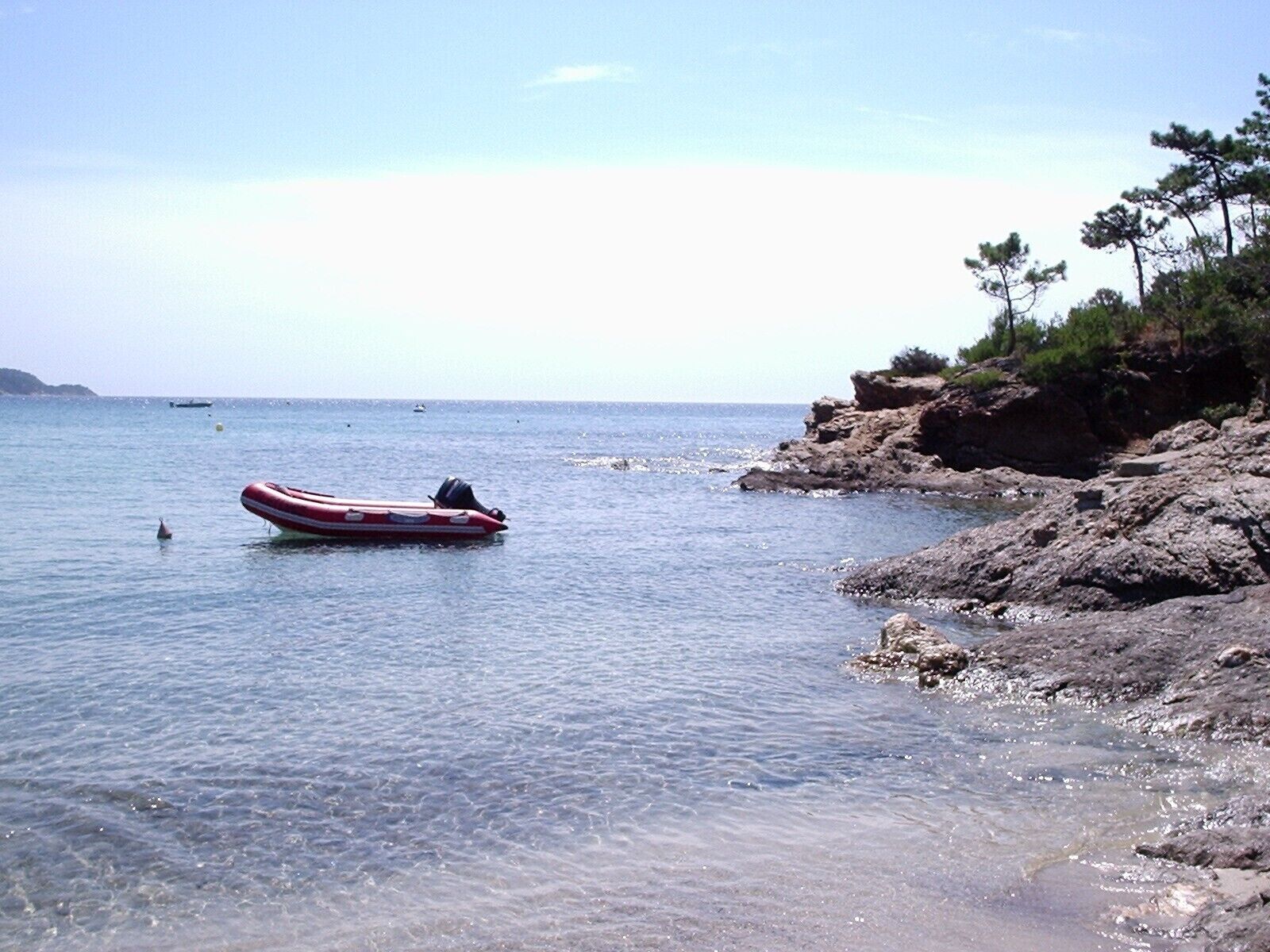 Ein klares Küstenbild mit einem kleinen Schlauchboot auf ruhigem Wasser, felsigem Ufer mit spärlicher Vegetation und Schiffen am Horizont.