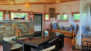 A room with a stone wall, wooden ceiling, and a bed with a canopy. There is a table with chairs and a bench with cushions.