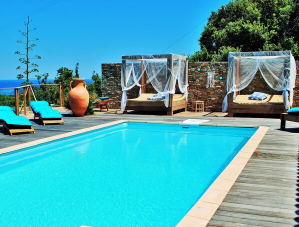 A clear blue pool with wooden deck, lounge chairs, and a stone wall with a view of the sea.