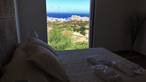 A bedroom with a large window offering a view of a coastal town and the sea.