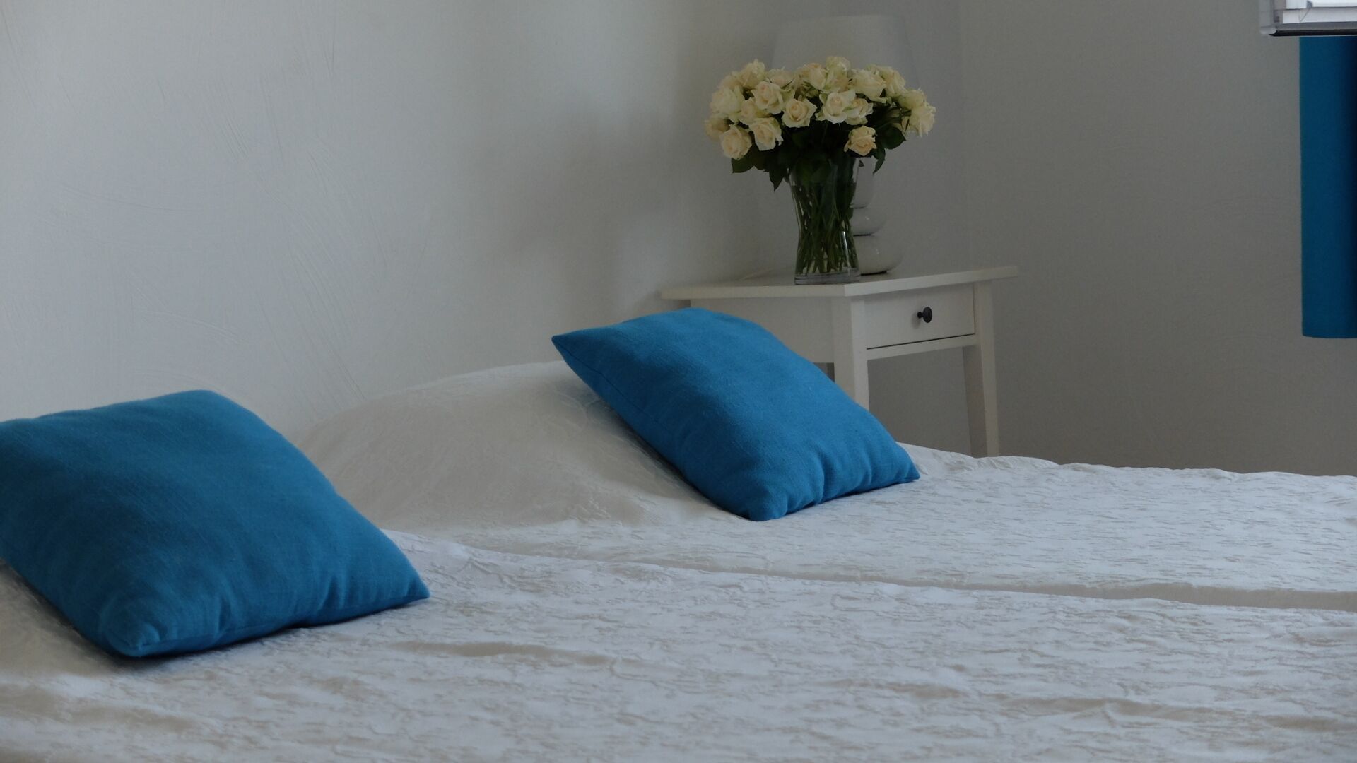 Ein Bett mit weißer Bettwäsche und zwei blauen Kissen, ein weißer Nachttisch mit Blumen in einer Vase und eine blaue Wand.