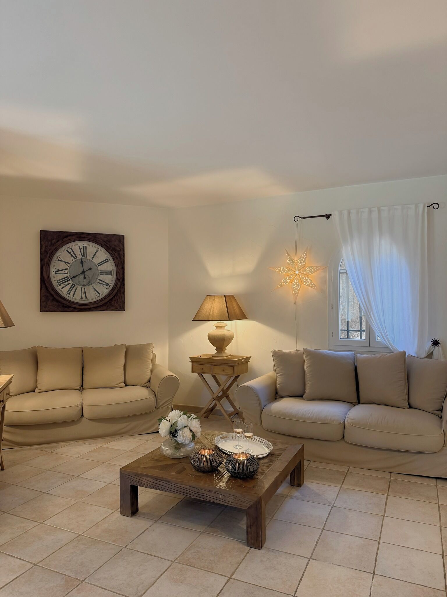 A living room with two beige sofas, a wooden coffee table, a wall clock, a lamp, and a star-shaped light fixture.