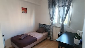 Une petite chambre avec un lit simple, un plancher en bois, un lit recouvert d’une couvre-lit rose et violet, une tête de lit noire, une petite table sur laquelle est posée une plante en pot et une fenêtre ornée de rideaux à motifs.