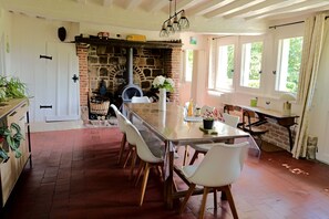 A dining area with a long table, white chairs, a brick fireplace, and a view of greenery outside.