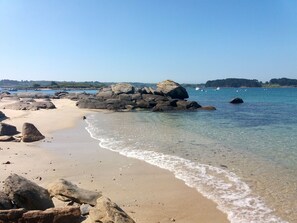 Une plage avec de gros rochers, une eau claire et des bateaux visibles au loin.