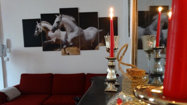 Un salón con un sofá rojo, un florero de vidrio con una vela roja y una pared con tres cuadros de caballos.