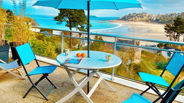 Ein Balkon mit Tisch, Stühlen und Sonnenschirm mit Blick auf einen Strand und eine Küstenstadt.