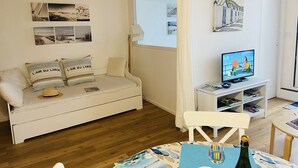 Ein gemütliches Wohnzimmer mit einem weißen Sofa, einem Fernseher und einem kleinen Tisch mit einer Flasche und Gläsern.