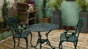 Una terraza de madera con una mesa redonda de metal y cuatro sillas, rodeada de macetas con plantas y una estatua de piedra.