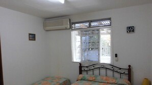 A bedroom with two single beds, a window with a view, an air conditioner, and a small bedside table with a lamp.