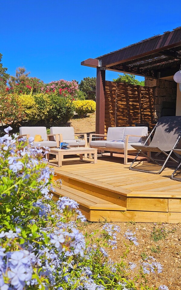Een terras met houten vloer, witte met kussens beklede stoelen en een pergola.