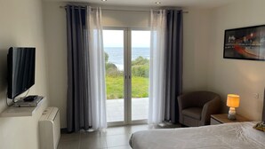 Una camera d'albergo con un letto, una sedia, una scrivania con una lampada, una TV e vista mare tra le tende.