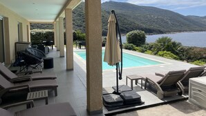 Un'area esterna coperta con poltrone relax, una piscina e vista sul mare e sulle montagne.
