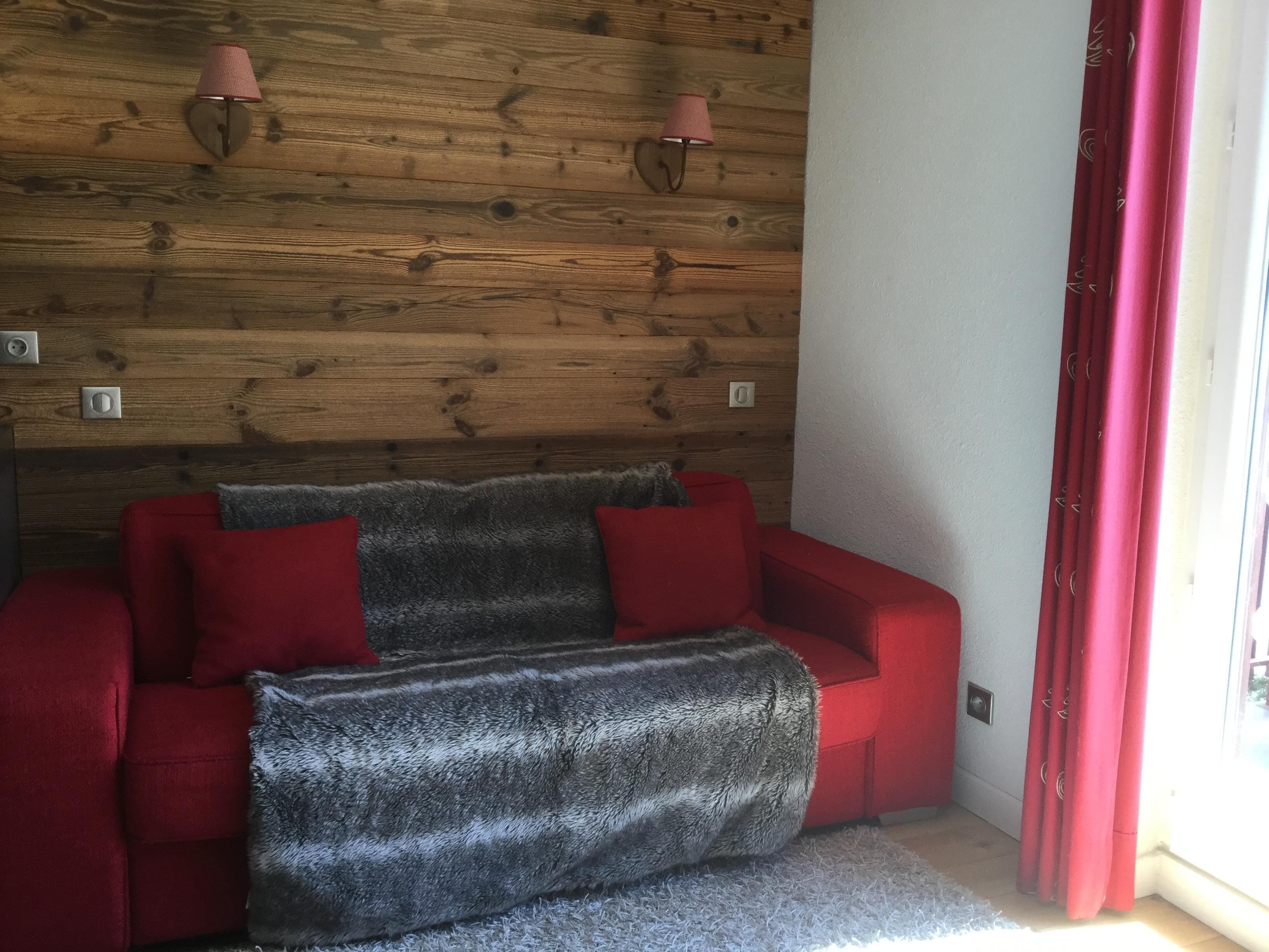 Une pièce chaleureuse avec un mur en bois, un canapé rouge, un plaid gris et des rideaux rouges.