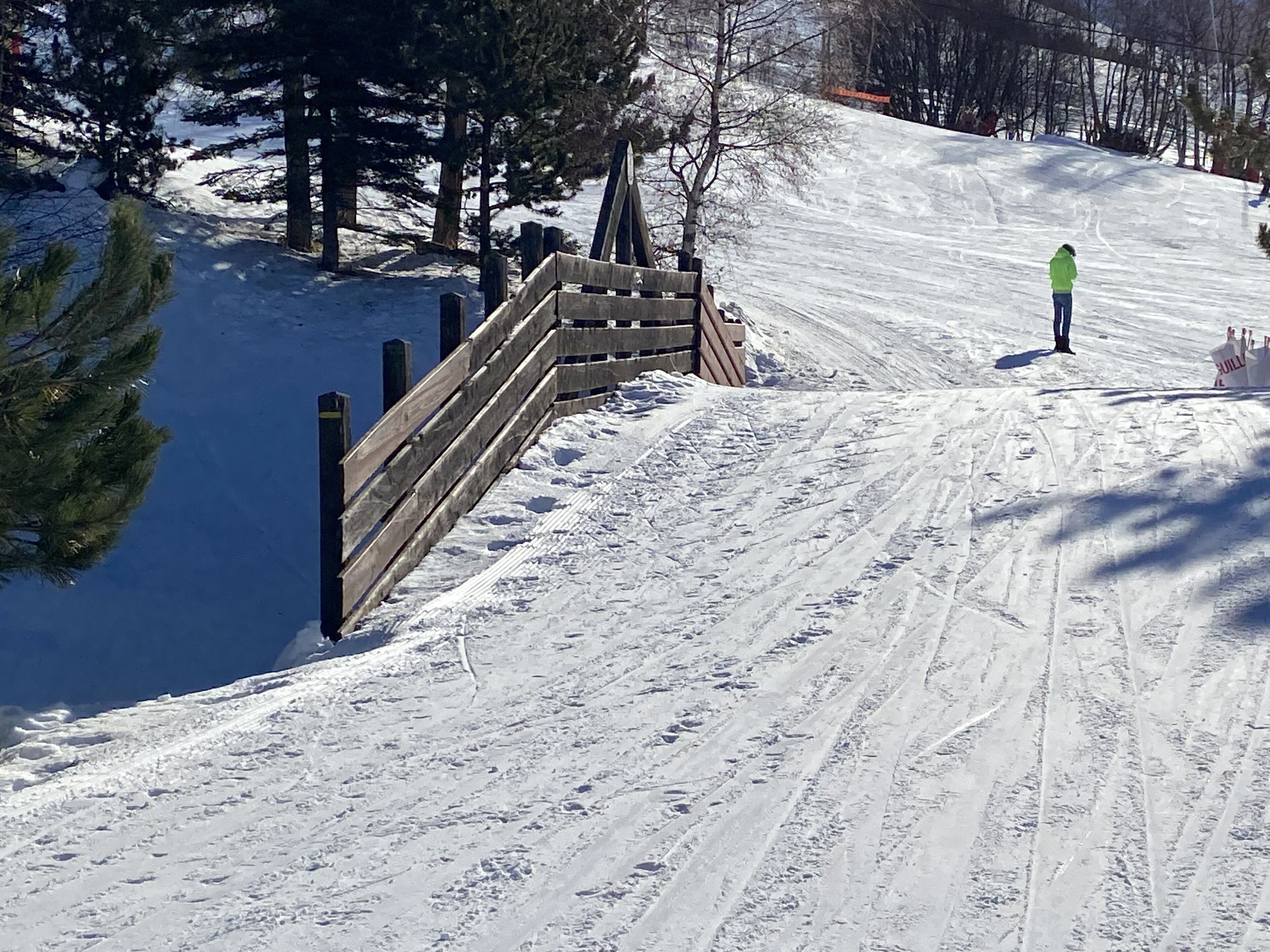 Pista de esquí nevada con una valla de madera y una persona con una chaqueta verde de pie a la derecha.