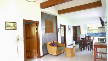 A spacious living area with a sofa, dining table, and chairs, a kitchen in the background, and a fan in the corner.
