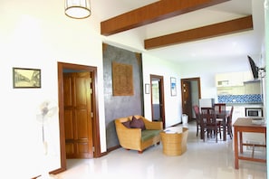 A spacious living area with a sofa, dining table, and chairs, a kitchen in the background, and a fan in the corner.