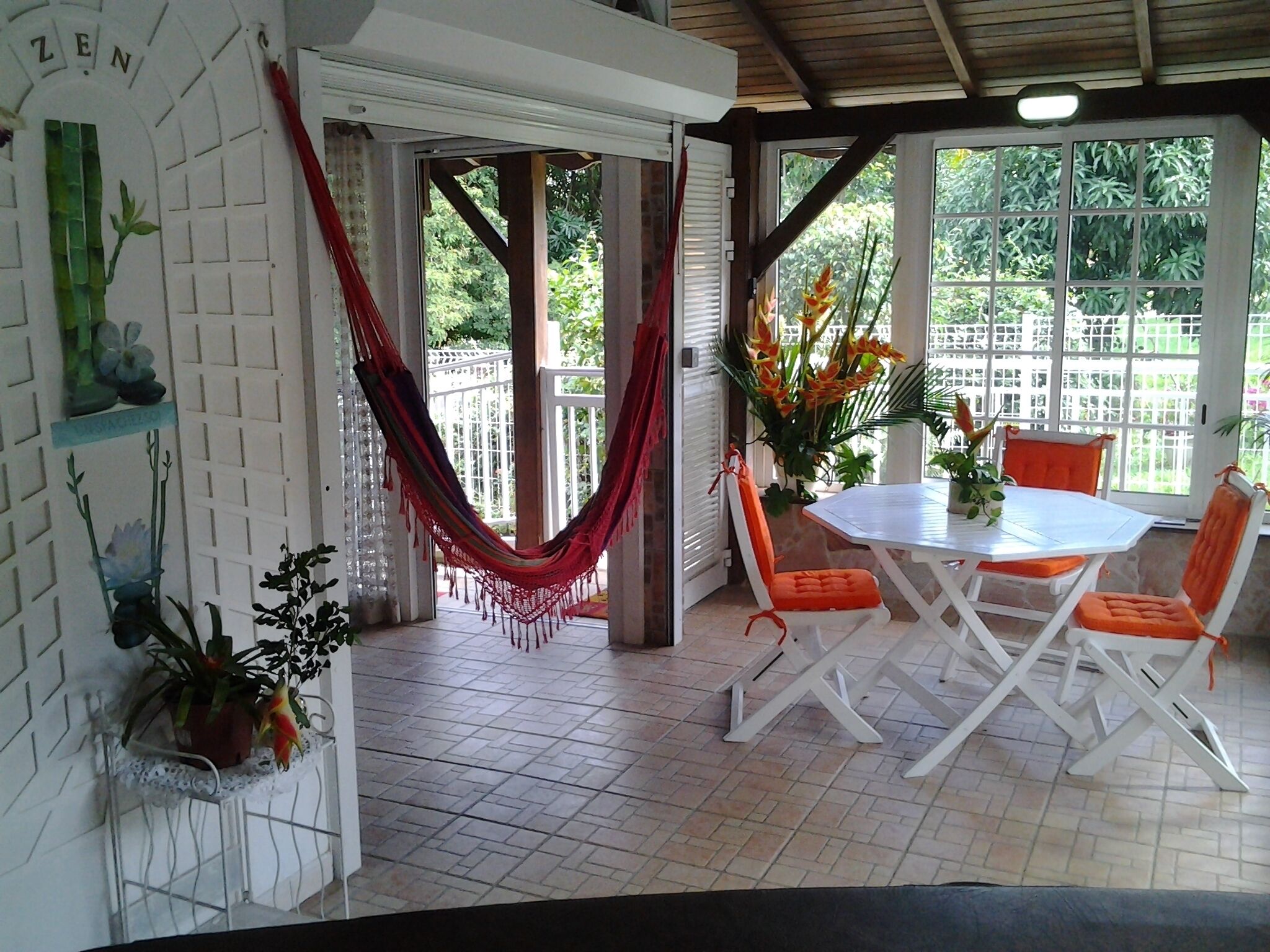 Une terrasse couverte avec une table blanche et des chaises orange, entourée de plantes en pot et d’un hamac.
