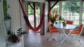 Une terrasse couverte avec une table blanche et des chaises orange, entourée de plantes en pot et d’un hamac.