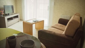 Una sala de estar con un sofá marrón, una mesa de centro de vidrio, un televisor sobre un mueble de madera y una mesa de comedor con cuencos.