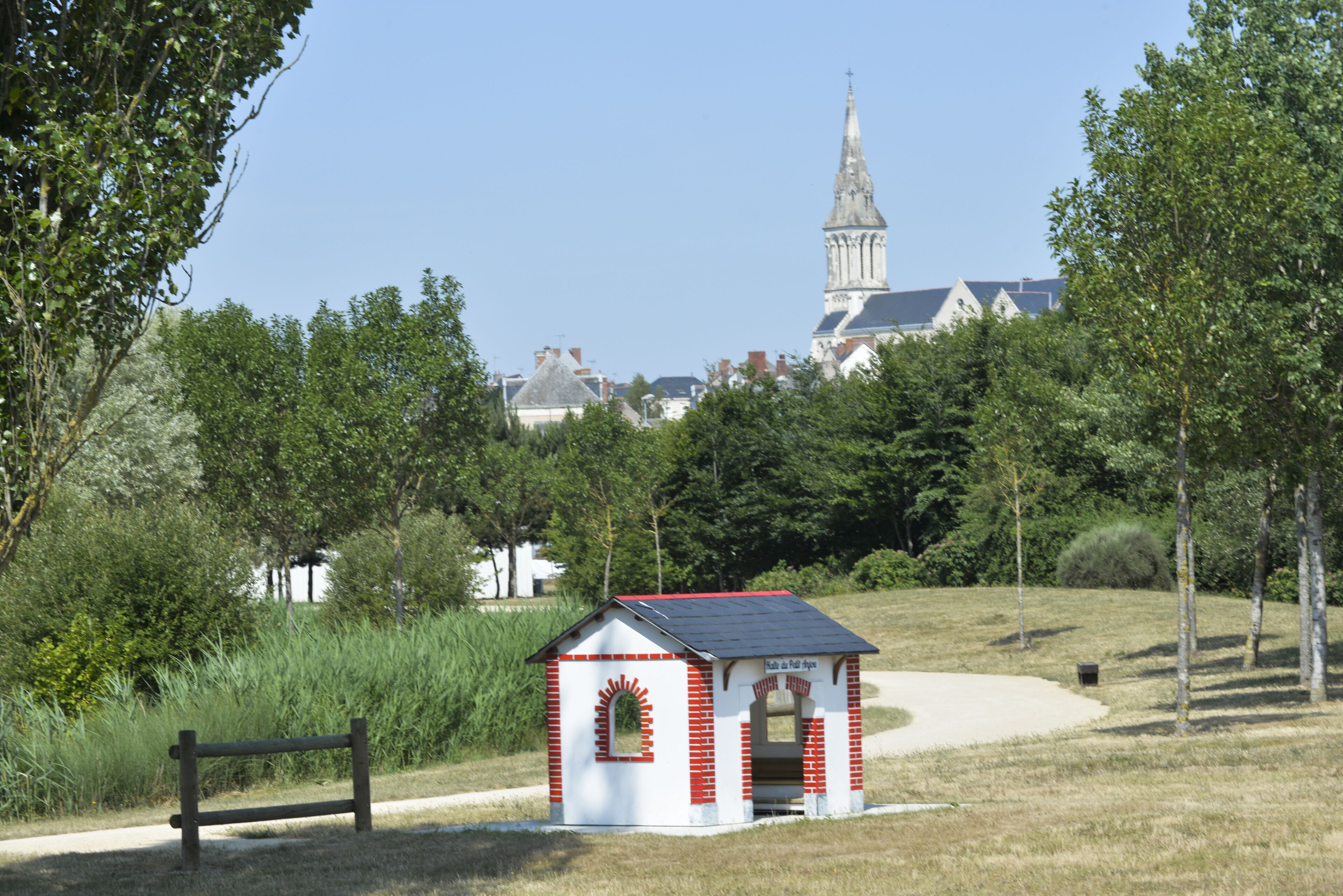 Una pequeña estructura roja y blanca con techo inclinado, rodeada de hierba y árboles, y una iglesia al fondo.