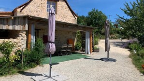 A stone house with a sloped roof, a covered patio area with chairs, and a gravel path leading to the house. - Charming cottage, private turquoise pool, PLAZAC-prox. treasures of Périgord (Plazac)