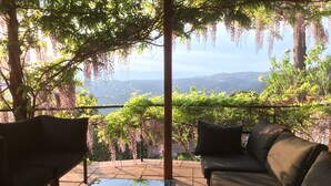 An outdoor seating area with a glass table and two sofas, surrounded by greenery and a view of distant hills.