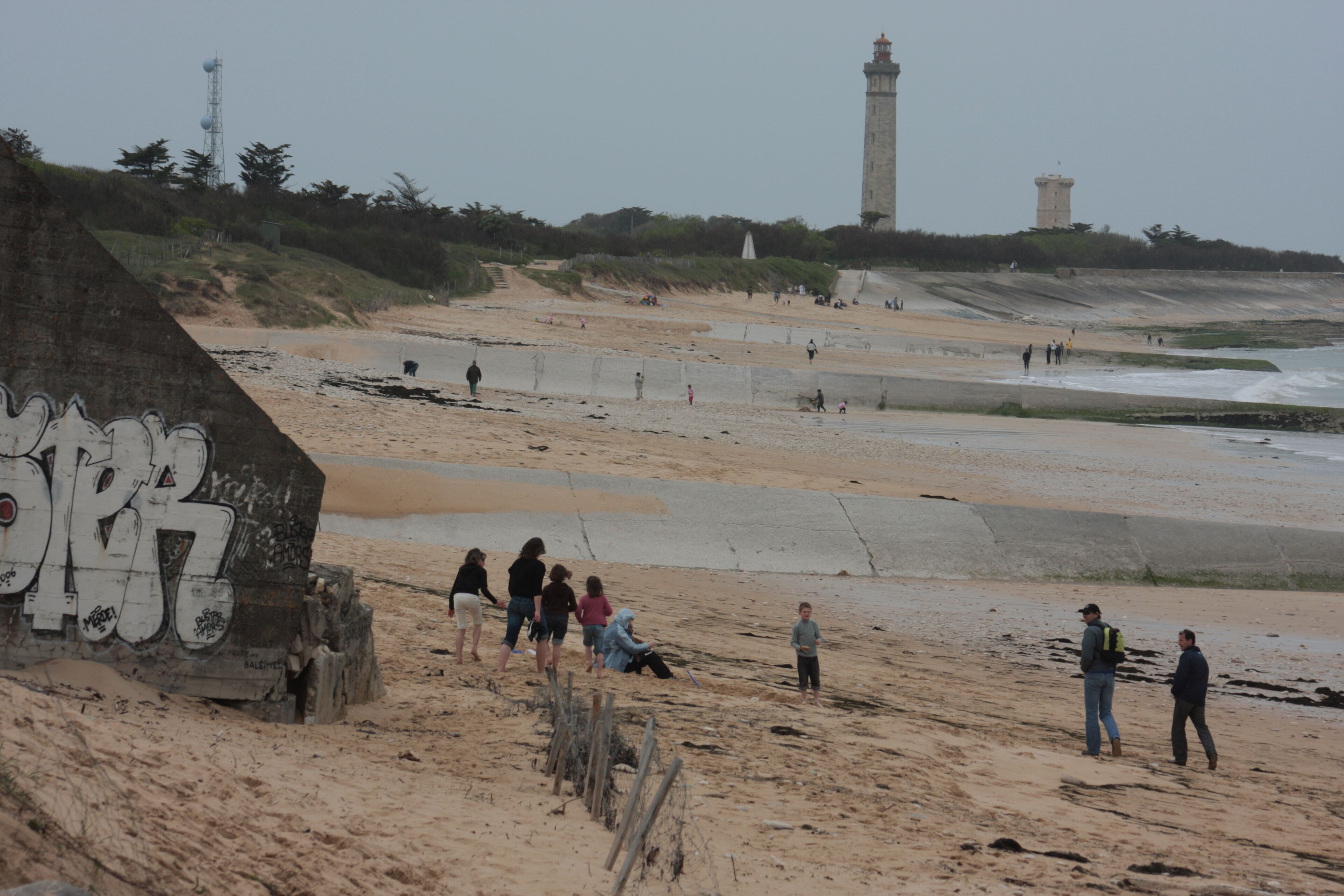 Ein Strand mit einem Leuchtturm im Hintergrund, sandigem Gelände und einer großen, mit Graffiti beschrifteten Struktur.