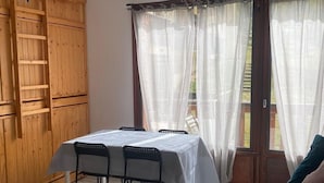 A room with a wooden wardrobe, a table with chairs, a bed with pillows, and a window with sheer curtains.