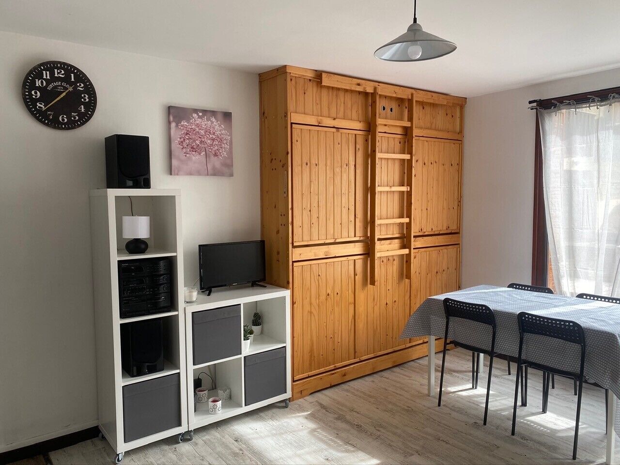 Une pièce comprenant une armoire en bois, un meuble TV blanc, une table à manger avec des chaises et une horloge au mur.