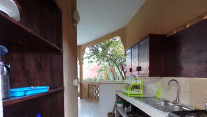 Cocina compacta con armarios de madera, estufa a gas y un lavamanos pequeño. Hay estantes con utensilios de cocina y vistas al exterior.