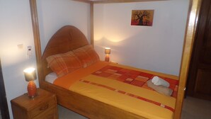 Una cama con dosel y tablero de madera, una manta escocesa y una colcha. Hay dos lámparas de noche y una mesita de noche de madera con una lámpara. En la pared, un cuadro de un árbol.