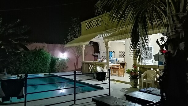 Un área de piscina nocturna con un espacio de estar cubierto, una cocina visible y una piscina con valla de seguridad.