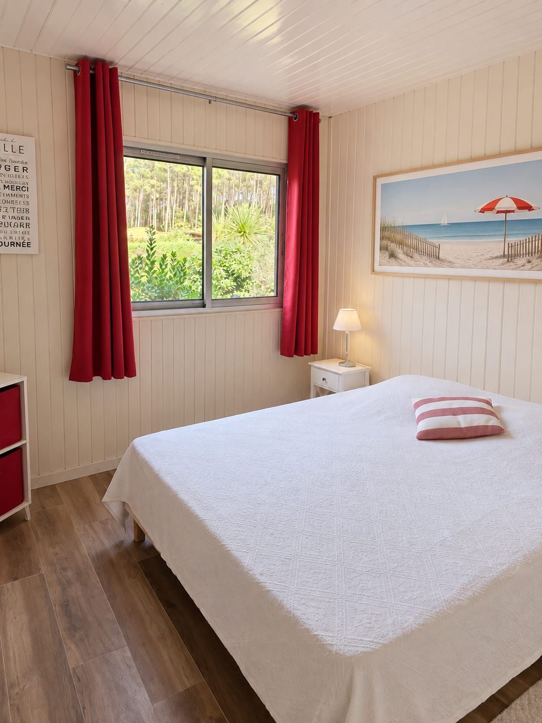 Una camera da letto con un letto, un comodino, una lampada, una finestra con tende, un cartello appeso al muro e un quadro raffigurante una spiaggia.