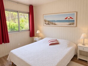 Un dormitorio con cama, mesita de noche, lámpara, ventana con cortinas y un cuadro con temática de playa en la pared.