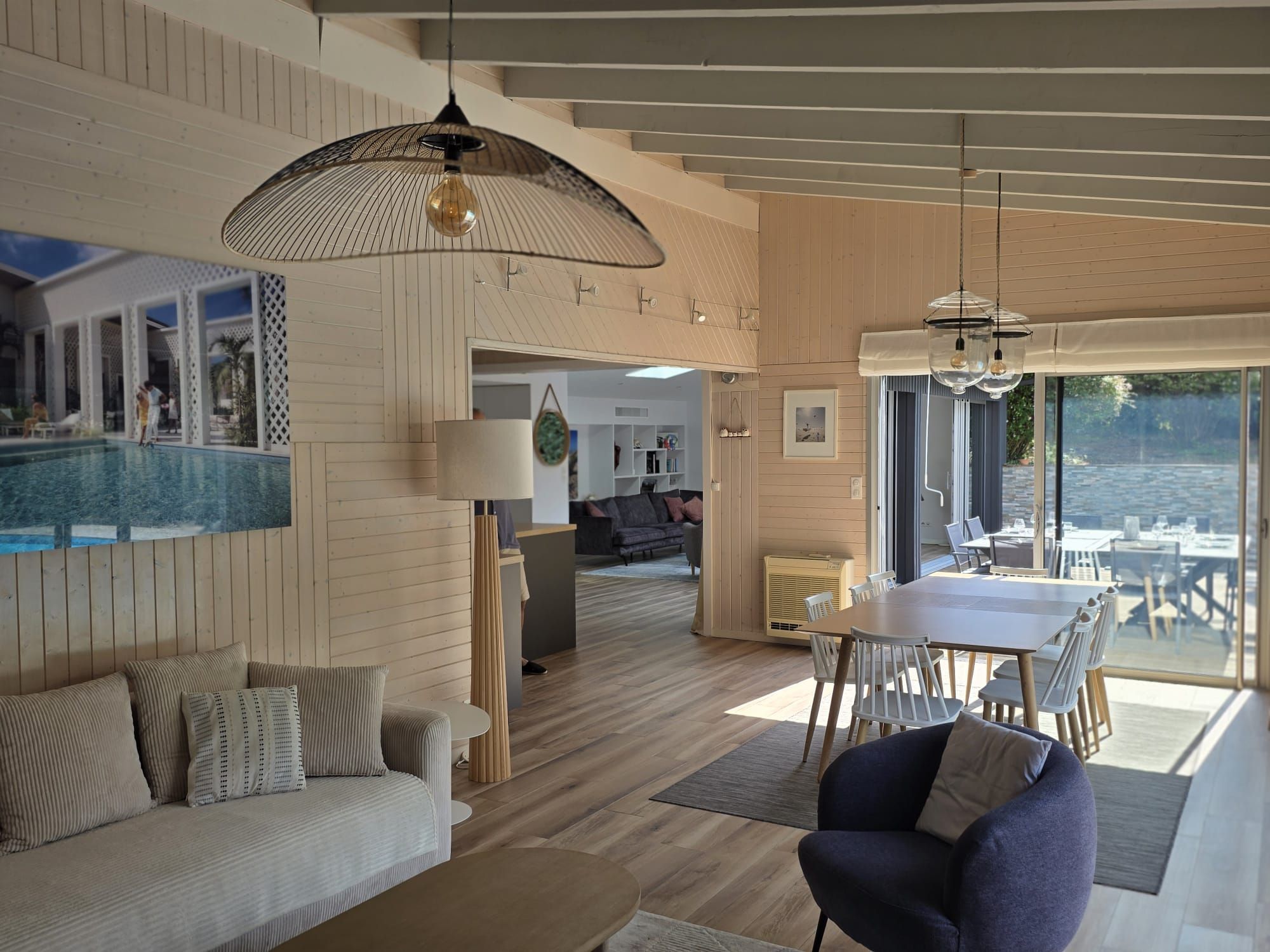 Un interior moderno con piso de madera, un área de comedor con sillas blancas, un sofá con cojines y un ventanal con vista al agua.