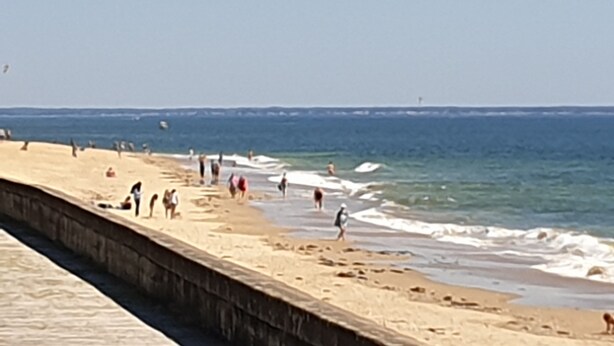 A beach with a concrete walkway along the water's edge, people walking and standing, and a clear sky.