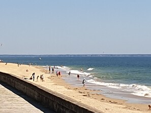 A beach with a concrete walkway along the water's edge, people walking and standing, and a clear sky.