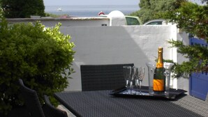 A table with a bottle of champagne and glasses, set against a backdrop of a white wall and a view of the sea.