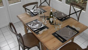 A dining table set with plates, cutlery, glasses, and a bottle, surrounded by chairs in a room with windows and a view of greenery.